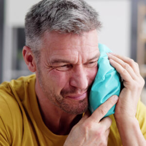 Middle-aged man with gray hair wincing while holding a teal ice pack to his cheek/jaw.