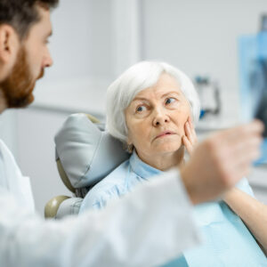 Elderly woman with white hair sitting in a dental chair, looking concerned while a male dentist shows her an X-ray.