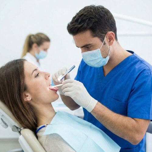 A male dentist in blue scrubs and a mask performing a thorough oral examination on a patient.